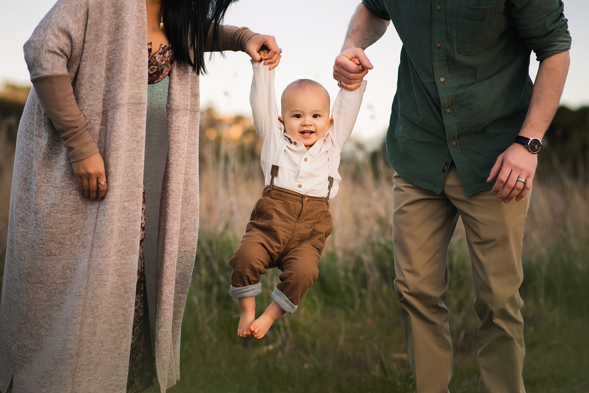 Mom and dad holding their baby while standing in a golden field at sunset, smiling and cuddling during a family photoshoot.